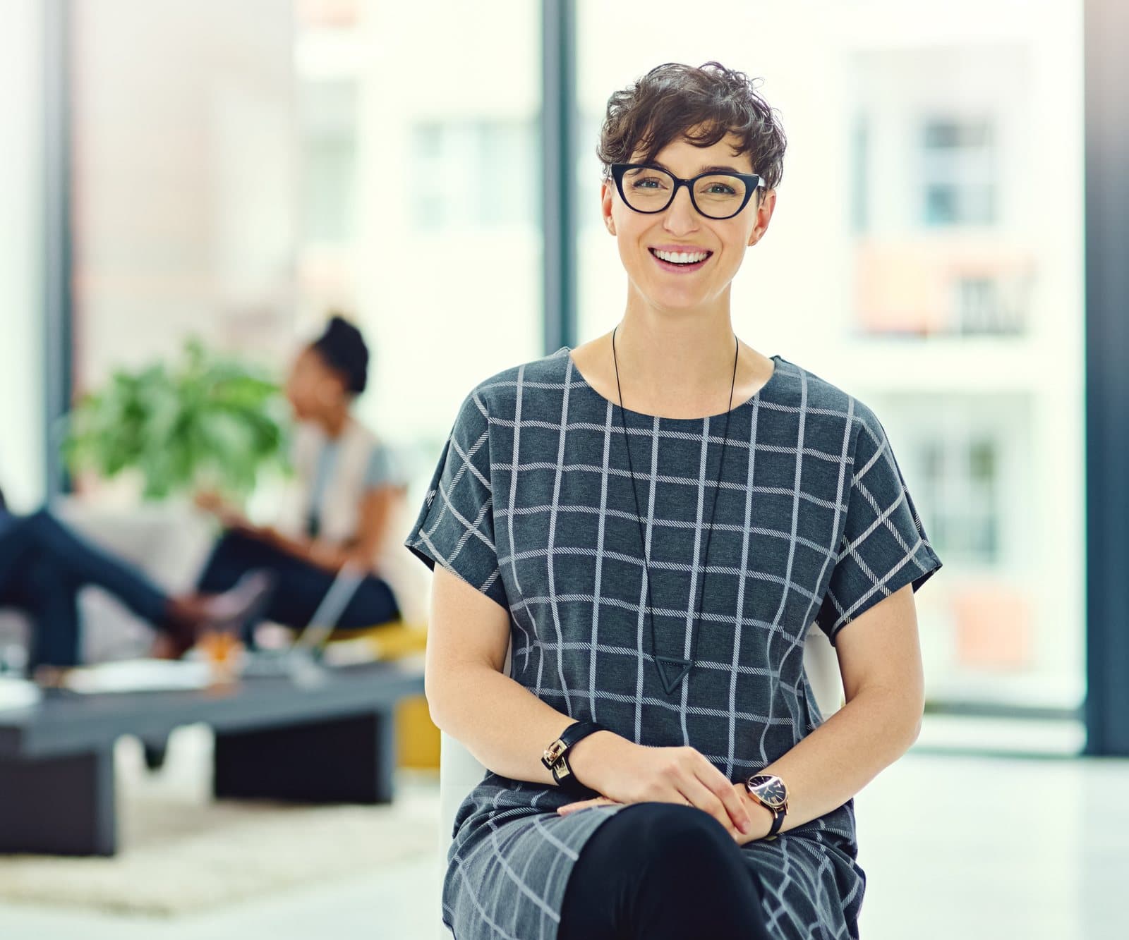 A smiling woman in glasses sits in a bright office while colleagues talk behind her