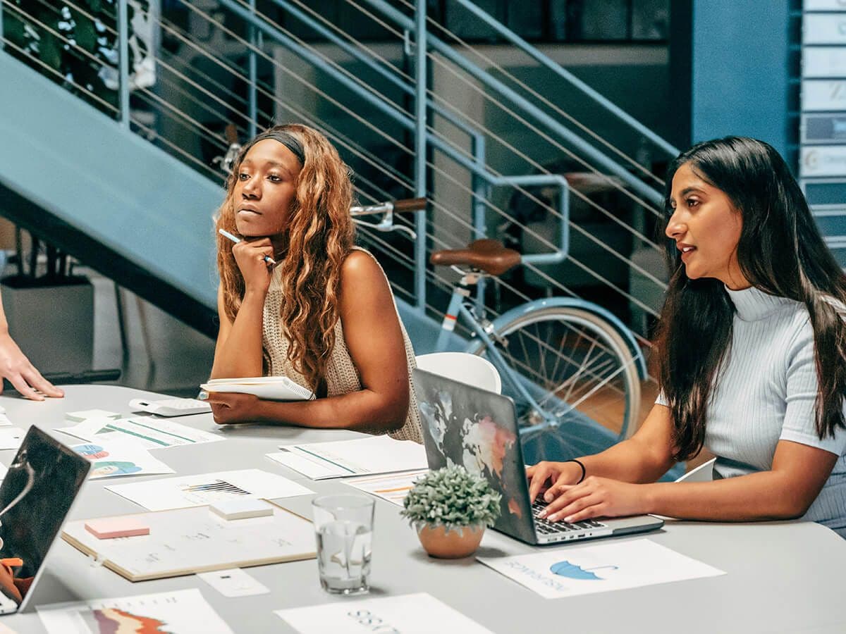 Two female employees sitting at a table in the open space of an office. They're on laptops an writing in notebooks at mentorship culture meeting.
