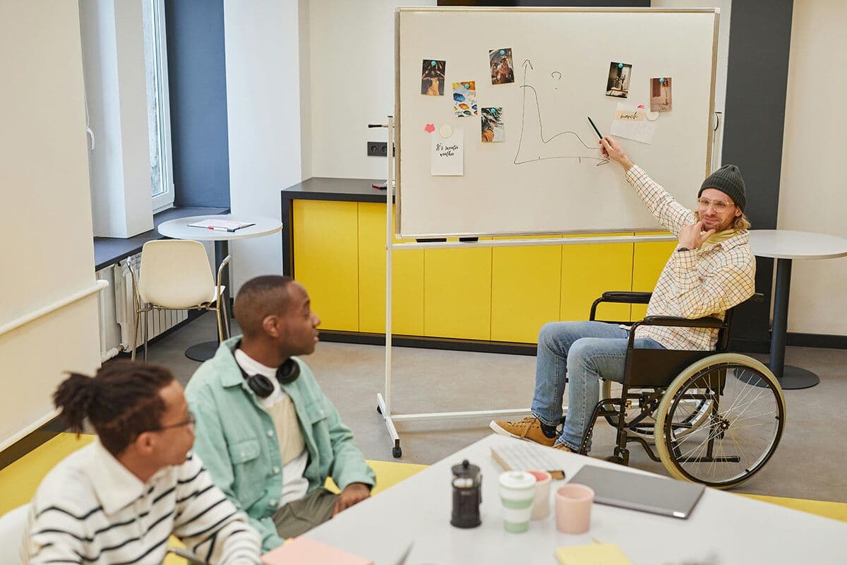 Man in a wheelchair points to a whiteboard in an office as two other men look at the board. They discuss servant leadership styles.