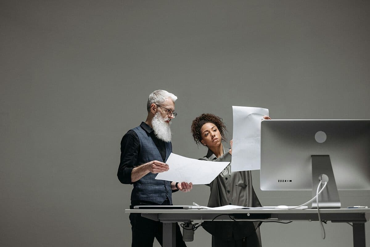 two employees standing at a desk and computer looking at pieces of paper